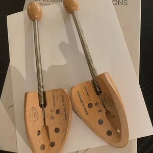 May include: Two wooden shoe trees with spring-loaded mechanisms. The shoe trees are made in Germany and have a size of 4-5. The shoe trees are on a white surface with the word "Calder" written in black ink.