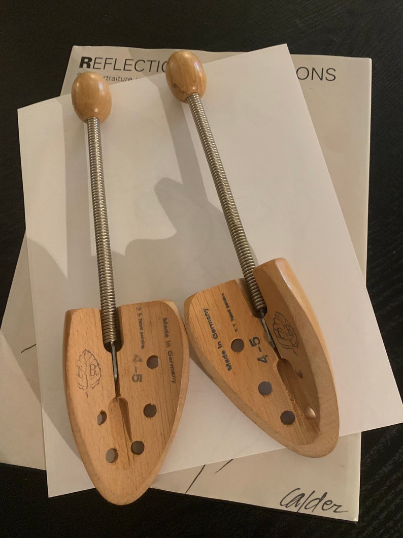May include: Two wooden shoe trees with spring-loaded mechanisms. The shoe trees are made in Germany and have a size of 4-5. The shoe trees are on a white surface with the word "Calder" written in black ink.