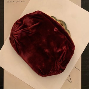 May include: A burgundy velvet clutch purse with a gold metal clasp and decorative embellishments. The purse is sitting on a white piece of paper with the text "REFLECTION Self-Portraiture in the Twentieth Century" and "Calder" written on it.