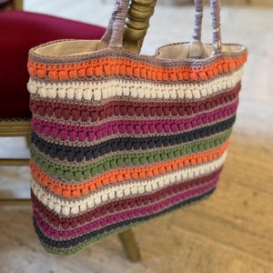May include: A colorful, striped crochet tote bag with a neutral interior lining. The bag features a variety of colors, including orange, white, green, and purple. The handles are a light brown color. The bag is sitting on a gold chair.