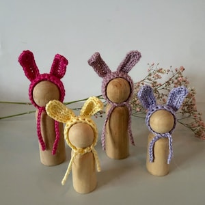 May include: Four wooden peg dolls wearing crocheted bunny hats in pink, yellow, lavender, and purple. Each doll has a different coloured hat with matching ties. The dolls are standing upright, and the background is a light colour.
