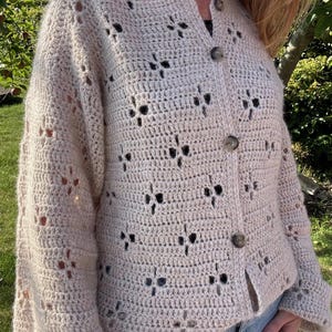 Op de afbeelding: Een beige gehaakt vest met open weefsel en een knoopsluiting. Het vest heeft een bloemenpatroon en lange mouwen. De knopen zijn in bijpassende beige kleur. Het kledingstuk wordt gedragen over een donkere top en een blauwe spijkerbroek.