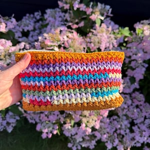 May include: A rectangular crochet pouch featuring horizontal stripes in vibrant colors including mustard yellow, purple, bright pink, turquoise, light blue, and sage green. The pouch has a white zipper closure along the top edge and a textured stitch pattern.