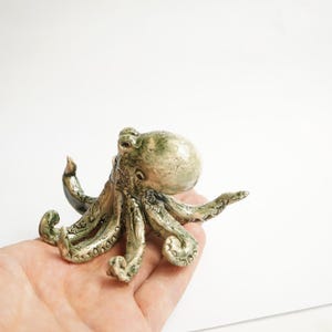 May include: A small ceramic octopus figurine with a green and brown glaze. The octopus has eight tentacles and is posed with its head raised.