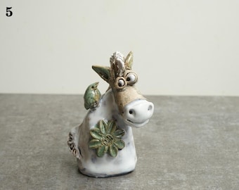 May include: A whimsical ceramic donkey figurine with a green bird perched on its back. The donkey has a white body, a tan face, and a green flower on its side. The figurine has a playful, cartoonish design.