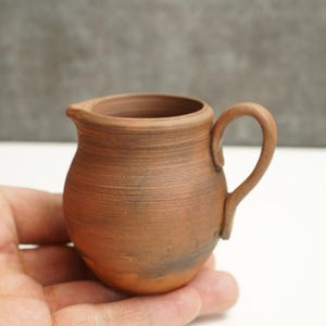 Può includere: Un piccolo brocca in ceramica marrone fatta a mano, con manico e beccuccio. La brocca ha un aspetto rustico e strutturato ed è tenuta nella mano di una persona. La brocca viene probabilmente utilizzata per servire liquidi.