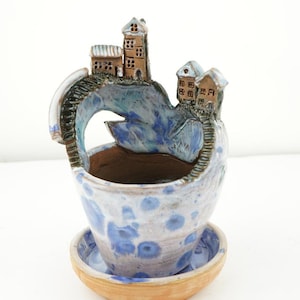 May include: A ceramic planter shaped like a cliff with houses on top. The planter is painted in shades of blue, white, and brown. It has a matching saucer.