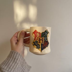 May include: Cream-coloured ceramic mug with the Hogwarts crest in red, green, blue, and yellow. The mug has a large handle and a slightly curved shape. The crest includes a lion, snake, badger, and eagle, with the letter "H" in the centre.