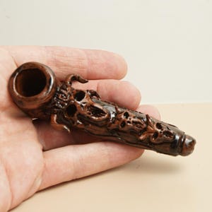 May include: A brown, carved wooden pipe with a detailed design. The pipe features a bowl, a stem, and intricate carvings along its surface. The design includes skull-like features and other decorative elements.