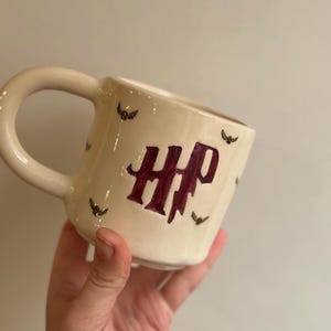 以下が含まれることがあります： クリーム色のセラミックマグカップ。大きなハンドルが付いています。「HP」の文字がバーガンディ色で描かれ、小さなコウモリのイラストが散りばめられています。ユニークな手作りのアイテムです。