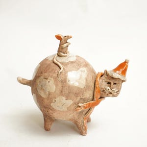 May include: A ceramic sculpture of a cat wearing a red and white hat and an orange scarf, with a small mouse perched on its back.