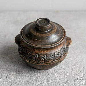 Puede incluir: Una olla de cerámica marrón oscuro con tapa. La olla tiene forma redonda con dos asas pequeñas a los lados. La parte inferior presenta un patrón floral decorativo. La tapa tiene un pequeño pomo en la parte superior.