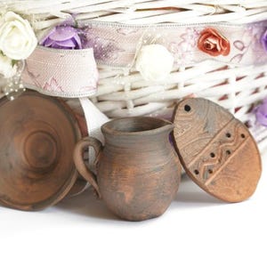 Pode incluir: Três pequenos vasos de barro marrom, um em forma de jarro, outro em forma de tigela e outro em forma de ovo. Os vasos estão sobre uma cesta de vime branca com uma fita rosa e branca e flores roxas e brancas.