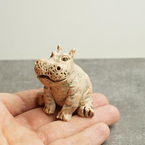 May include: A small, detailed figurine of a hippopotamus in a seated position. The sculpture is a light beige color with textured details, including the hippo's skin and facial features. The figurine is held in a hand.