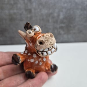 Peut inclure: Une figurine de cheval en céramique fantaisiste avec un large sourire. Le cheval est orange avec des taches blanches et des sabots et une crinière brun foncé. La figurine a de grands yeux expressifs et mesure environ 7.6 cm de haut.