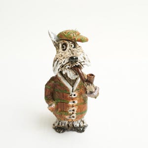Può includere: Una statuetta in ceramica di un cane che indossa un cappello verde e marrone e una giacca marrone. Il cane tiene una pipa in bocca.