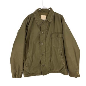 May include: Olive green jacket with a button-up front, two front pockets, and a collar. The jacket is hanging on a white hanger. The jacket has a label on the inside collar.