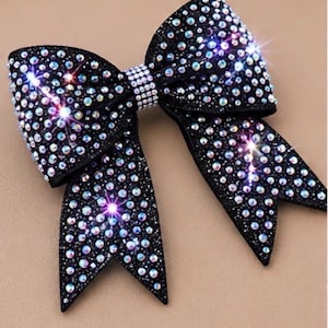 May include: A black bow adorned with numerous iridescent rhinestones. The bow features a central band of smaller, clear rhinestones, and the overall design is symmetrical with pointed ends. The rhinestones catch the light, creating a sparkling effect.