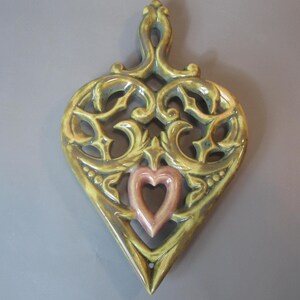 May include: A ceramic heart-shaped hot pad with an intricate design in shades of green and brown. The center of the heart features a smaller pink heart.