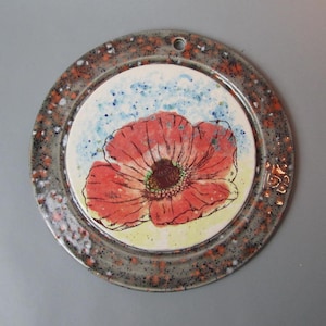 May include: Round ceramic wall hanging with a hand-painted red poppy flower design. The poppy is centered on a white background with blue speckles, surrounded by a speckled gray and orange rim. A small hole is at the top for hanging.