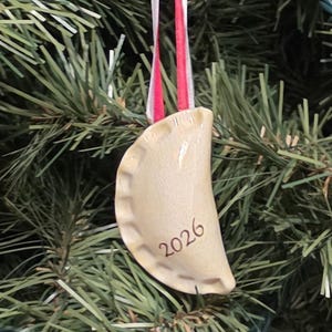 May include: A cream-colored ceramic dumpling ornament with a crimped edge and the year 2026 printed in dark brown text. The ornament is suspended by a thin red and white ribbon, featuring a smooth, glossy finish on its curved, crescent shape.