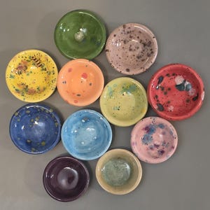 Può includere: Una collezione di undici piccole ciotole rotonde in ceramica di vari colori, tra cui verde, rosa, giallo, arancione, blu, rosso e viola. Alcune ciotole presentano motivi maculati, mentre una ciotola gialla mostra una piccola illustrazione di un'ape.