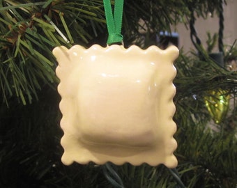 Può includere: Un ornamento di ceramica bianca a forma di raviolo con un nastro verde. L'ornamento è a forma di raviolo e ha un bordo smerlato.