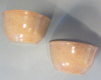 May include: Two ceramic bowls with a speckled, light orange glaze. The bowls are empty and sitting on a light gray surface.