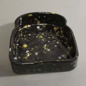 Peut inclure: Un plat en céramique noire en forme de tranche de pain avec un glaçage moucheté de blanc, jaune et noir.