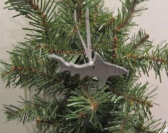 May include: A silver shark-shaped Christmas ornament hanging on a green Christmas tree. The ornament is made of a shiny, metallic material and is suspended by a silver ribbon. The shark has detailed fins and a tail.