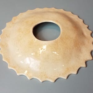May include: A cream-coloured ceramic lampshade with a scalloped edge and a large central hole. The lampshade has a slightly textured surface.