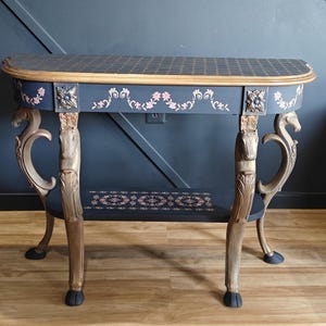 May include: A navy blue console table with gold accents and floral details. The table has a patterned top, ornate legs, and a lower shelf. The legs are carved with horse head designs. The table is a decorative furniture piece.