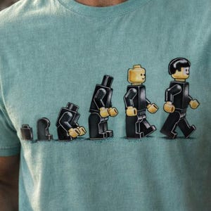 May include: A light blue t-shirt featuring a graphic of Lego figures in a walking sequence. The person in the image is holding a trowel. The background shows an archaeological site.