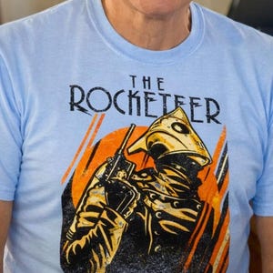 May include: A man wearing a light blue t-shirt with the text "THE ROCKETEER" and a graphic of a rocket-powered character. He is smiling and wearing black-framed glasses. The background includes a sign that says "SUNSETS".