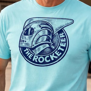 May include: Light blue t-shirt featuring a circular graphic with the text "The Rocketeer" and a stylized helmet design in navy blue. The shirt is short-sleeved and worn by a person wearing sunglasses and khaki pants.