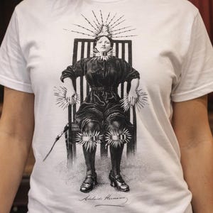 May include: White t-shirt featuring a black and white illustration of a woman seated in a chair, with a crown and radiating lines. The shirt is worn by a person in front of a stage with red curtains.