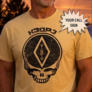 Könnte beinhalten: Senfgelbes T-Shirt mit einem schwarzen, abgenutzten Grafikdesign, das einen Totenkopf, eine Raute und eine Antenne zeigt. Der Text "K3QPJ" befindet sich über der Raute. Eine Sprechblase sagt "YOUR CALL SIGN".