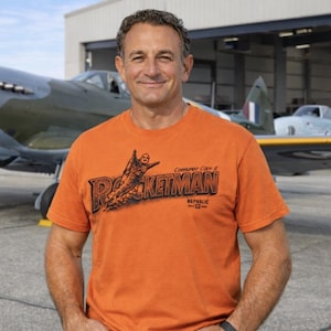 Puede incluir: Camiseta naranja con el texto "Rocketman" y un gráfico de una figura con un jetpack. La camiseta la lleva una persona de pie frente a un avión antiguo. La camiseta también tiene el texto "Commando Cody is Republic -12-".