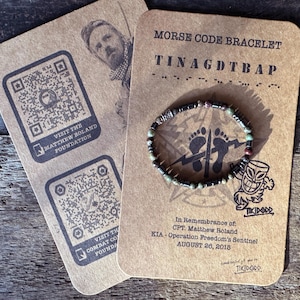 Puede incluir: Una tarjeta marrón con un brazalete de código Morse en blanco y negro con cuentas verdes y rojas. La tarjeta tiene el texto "MORSE CODE BRACELET" y "TINAGDTBAP" en negro. La tarjeta también tiene el texto "En memoria de: CPT. Matthew Roland KIA - Operación Freedom's Sentinel 26 de agosto de 2018" en negro.