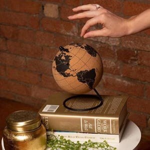 Peut inclure: Un globe terrestre en liège avec des continents noirs repose sur un support en métal noir, posé sur une pile de livres. Le globe mesure environ 15 cm de diamètre. Un pot doré avec un couvercle est au premier plan.