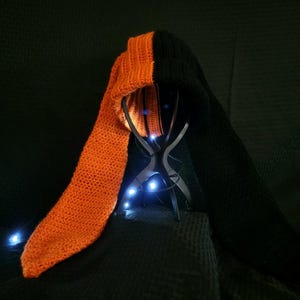 Könnte beinhalten: Handgefertigtes Häkel-Hoodie- und Schal-Set. Die Kapuze ist schwarz und der Schal orange. Das Set wird auf einem Metallständer mit blauen Lichtern präsentiert. Die Häkelarbeit scheint engmaschig zu sein, was dem Set ein strukturiertes Aussehen verleiht.