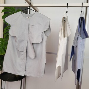 May include: Three sleeveless tops hanging on a rack. The first is light gray with a unique wavy design around the shoulders and a small pocket. The second is white, and the third is light blue with a dark blue trim.