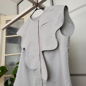 May include: A light gray sleeveless top with a unique, asymmetrical design. The top features a large, wavy collar and a button-down front. The fabric appears to be a textured linen or cotton blend. The garment is hanging on a black hanger.