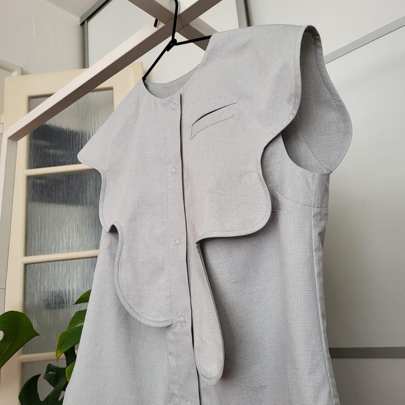 May include: A light gray sleeveless top with a unique, asymmetrical design. The top features a large, wavy collar and a button-down front. The fabric appears to be a textured linen or cotton blend. The garment is hanging on a black hanger.