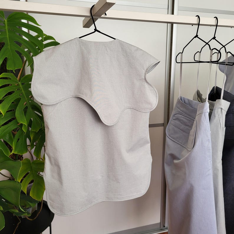 May include: A light gray top with a unique wavy collar, hanging on a black hanger. The top is made of a soft fabric and has a simple, minimalist design. Other clothing items are visible on hangers in the background.