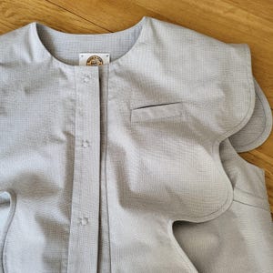 May include: A light gray, sleeveless garment with a rounded neckline and scalloped edges. The front features a button closure, a small pocket, and a label near the collar. The fabric has a subtle textured pattern.