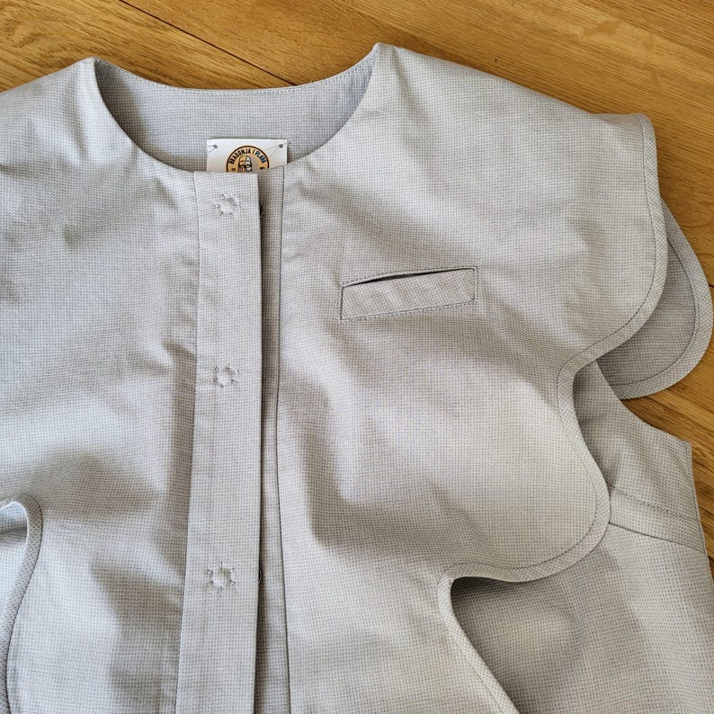 May include: A light gray, sleeveless garment with a rounded neckline and scalloped edges. The front features a button closure, a small pocket, and a label near the collar. The fabric has a subtle textured pattern.