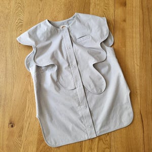 May include: A light gray button-down shirt with a unique, wavy collar design. The shirt features a small chest pocket and a rounded hem. The fabric appears to be a lightweight cotton or linen, suitable for warm weather. The shirt is unadorned, with a minimalist aesthetic.