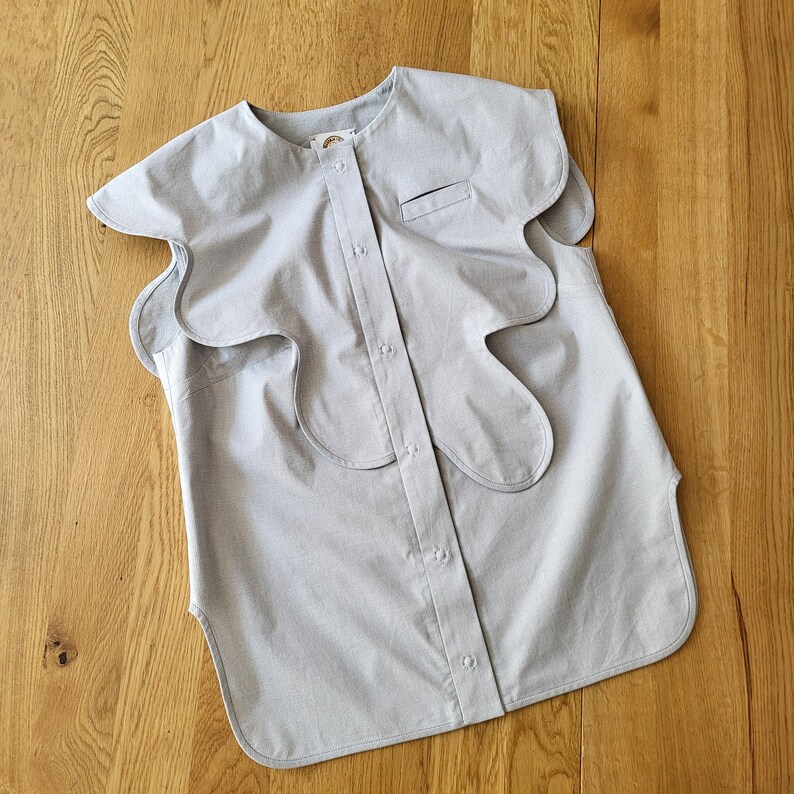 May include: A light gray button-down shirt with a unique, wavy collar design. The shirt features a small chest pocket and a rounded hem. The fabric appears to be a lightweight cotton or linen, suitable for warm weather. The shirt is unadorned, with a minimalist aesthetic.