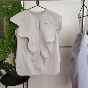 May include: A light gray sleeveless top with a unique, wavy collar design. The top has a button-up front, a small chest pocket, and is hanging on a black hanger. Another similar top is visible on the right.